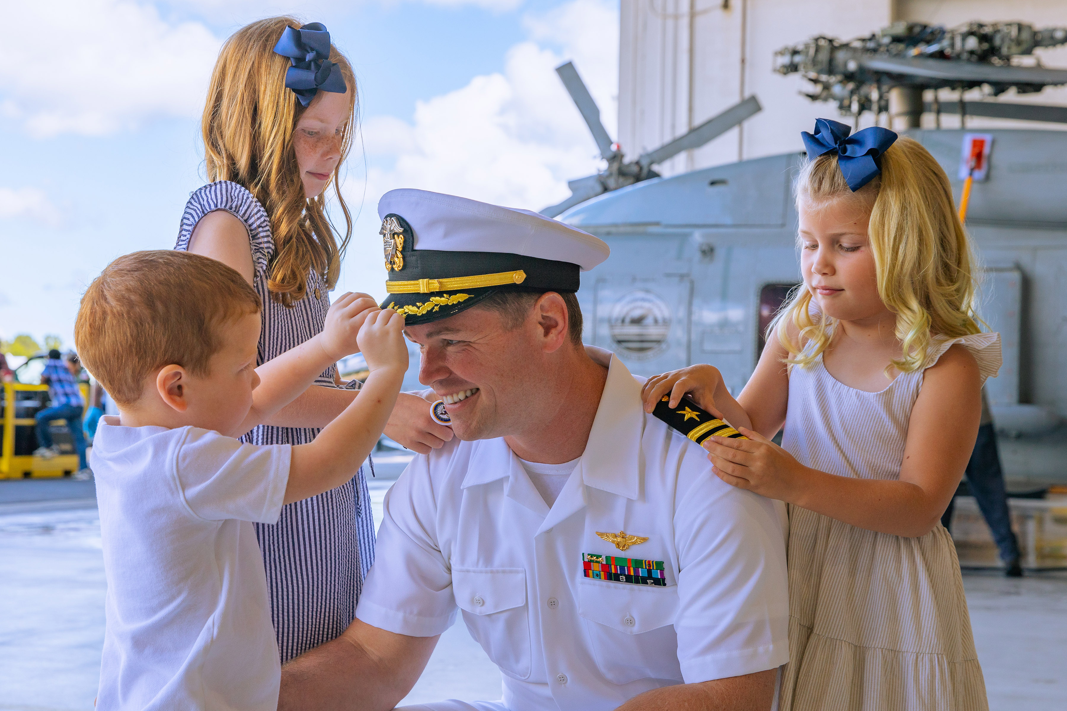 A Navy servicemember in dress white uniform kneeling with his three yound children